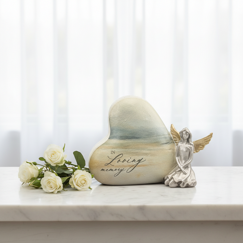 Heart-shaped memorial stone with a painted ocean scene and the words 'In Loving Memory' on a white marble surface, styled with white roses and a small kneeling angel figurine.