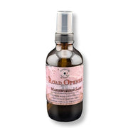 An amber glass spray bottle with a silver pump dispenser and a pink label reading 'Road Openers' and 'Manifestation Lane ', shown on a white background.