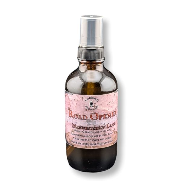 An amber glass spray bottle with a silver pump dispenser and a pink label reading 'Road Openers' and 'Manifestation Lane ', shown on a white background.