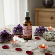 A bottle of Road Openers Manifestation Lane Scented Spray stands on a light tabletop surrounded by purple amethyst clusters, white crystal points, a bundle of white sage, and lit tealight candles. The product label is pink and readable, with the brand name Road Openers visible.