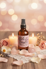 An amber glass spray bottle labeled 'Love/Attraction' sits on a wooden surface surrounded by pink rose petals, soft-focus pink roses, lit candles, and a lace doily against a warm bokeh background.