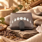 A black moon phase wallet on a piece of stone surrounded by herbs, rocks, beige materials and candles.