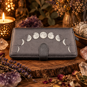 A dark gray leather wallet featuring a moon phase design on the front, displayed on a rustic wooden slice surrounded by crystals, dried flowers, and a lit candle.