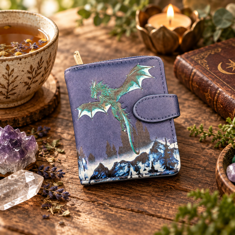 Small pouch with a dragon design on a purple background on a wooden table surrounded by a cup of tea, candle, crystals and a leather bound book.