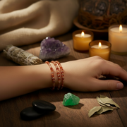 A close-up of a person's hand resting on a wooden surface, wearing three beaded orange-red bracelets. The scene is styled with candles, a smudge bundle, crystals, and black stones in a warm, atmospheric setting.