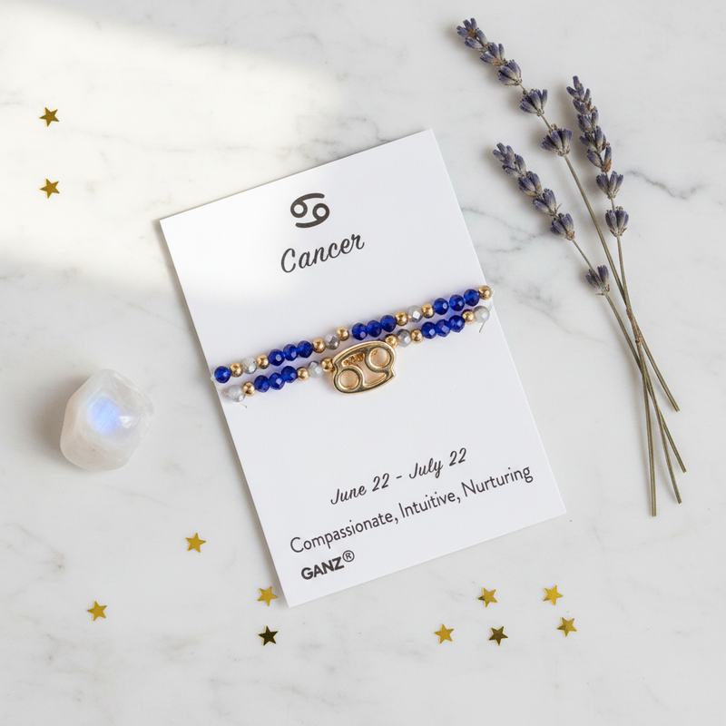 A Cancer zodiac bracelet by GANZ, featuring blue and gold beads with a gold Cancer symbol charm, displayed on its white card against a marble background with lavender and gold star decorations.