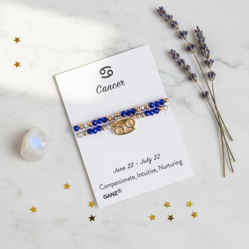 A Cancer zodiac bracelet by GANZ, featuring blue and gold beads with a gold Cancer symbol charm, displayed on its white card against a marble background with lavender and gold star decorations.