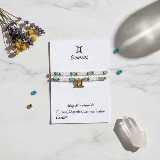 A Gemini zodiac-themed beaded bracelet , displayed on a white card. The card is styled on a white marble surface with dried lavender, small scattered beads, and a clear crystal.