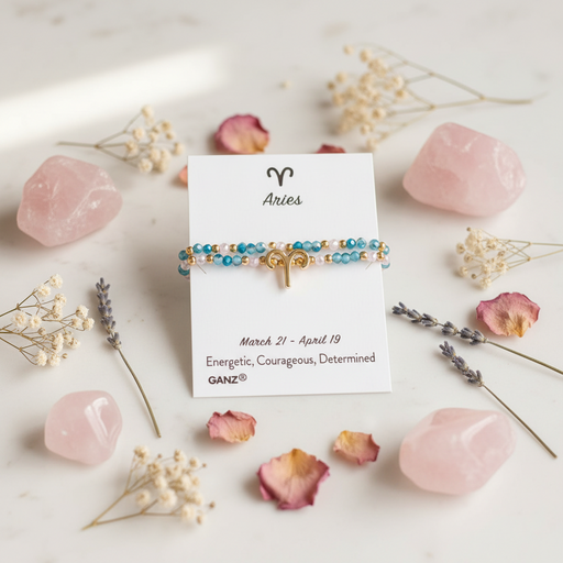 A beaded Aries astrology bracelet with blue, gold, and white beads and a small gold ram charm, displayed on a white card labeled 'Aries' with pink crystals and dried flowers around it.