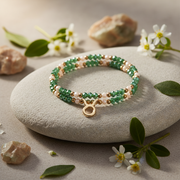 A beaded stretch bracelet with green and white beads and a gold-tone Taurus zodiac charm, displayed on a smooth light gray stone surrounded by small white flowers and leaves.
