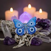 A decorative blue and white butterfly ornament with large circular eye-like motifs on its wings, styled on a dark surface with lit candles, crystals, and dried white sage in the background.