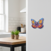 A colorful butterfly magnet  with vibrant blue, orange, yellow, and purple patterns, shown on a white refrigerator door in a blurred kitchen setting.