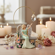 A decorative angel figurine seated on a bench, wearing a pale green dress and reaching toward a hanging teardrop crystal under a gold arched fixture. The scene is styled with lit candles, dried flowers, crystals, and soft background decor.