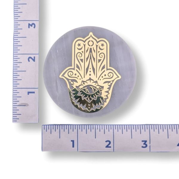 A selenite charge plate with a golden Hand Of Fatima design next to a ruler for scale on a white background
