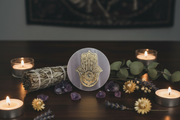 A  selenite charge plate with a golden Hand Of Fatima design, styled on a dark wooden table with burning candles, a bundle of dried herbs, purple crystals, and eucalyptus leaves.