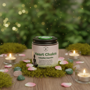 Candle jar labeled 'Heart Chakra' on a wooden surface with candles and stones, blurred greenery in the background