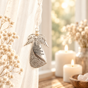 An engraved silver angel-shaped keepsake ornament hangs from a white ribbon in a softly lit home setting with white candles, dried flowers, and a sheer curtain.