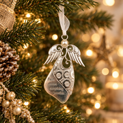 A silver angel-shaped Christmas ornament hanging from a white ribbon on a decorated Christmas tree with warm bokeh lights in the background. The ornament features floral patterns and the engraved text 'Faith makes everything possible'.