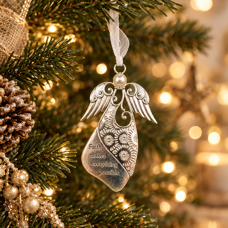 A silver angel-shaped Christmas ornament hanging from a white ribbon on a decorated Christmas tree with warm bokeh lights in the background. The ornament features floral patterns and the engraved text 'Faith makes everything possible'.