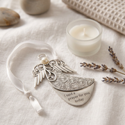Silver angel-shaped ornament with engraved details, a white pearl accent, and a white ribbon loop, resting on a textured cream linen surface next to a small white candle, dried lavender, and smooth stones.