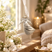 A silver angel-shaped hanging ornament with engraved text reading 'Angels are everywhere' and a small pearl detail, suspended by a sheer white ribbon in a softly lit room near a window with white flowers.