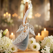 A silver angel-shaped hanging ornament with engraved wings and the words 'Let go and let God' on the body, suspended from a wooden stand with a white ribbon. The ornament is photographed in a softly blurred indoor setting with candles and white flowers around it.