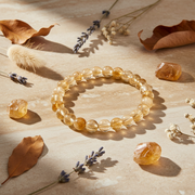 A beaded stretch bracelet made of translucent citrine-yellow crystal round beads, arranged on a warm beige surface with dried leaves, lavender sprigs, and small polished stones around it.