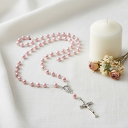 A pink pearl rosary with a silver crucifix and centerpiece, arranged on a white fabric surface next to a white candle and small dried flowers.