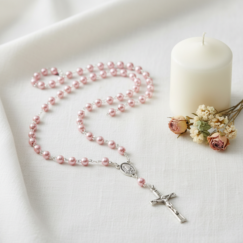 A pink pearl rosary with a silver crucifix and centerpiece, arranged on a white fabric surface next to a white candle and small dried flowers.