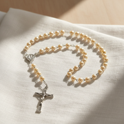 A beaded rosary with cream-colored pearls and a silver-tone crucifix pendant, arranged on a textured off-white fabric under warm natural sunlight.