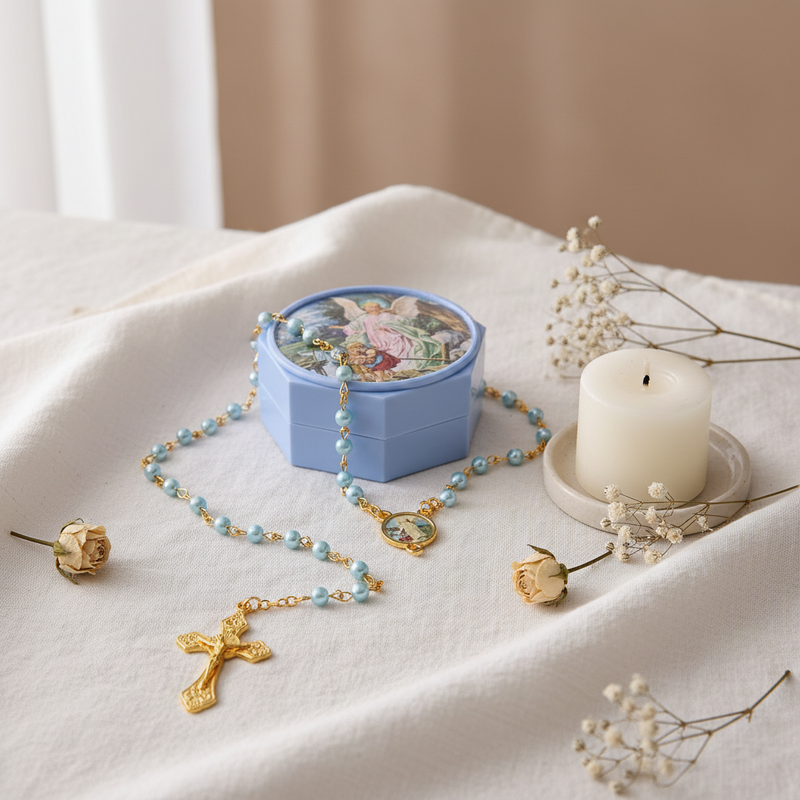 A light blue hexagonal rosary box with a colorful illustration on the lid, shown with a beaded rosary featuring light blue beads and gold-tone cross and heart pendants. The rosary is styled on a draped white fabric surface alongside a white candle, dried rose buds, and baby's breath flowers.
