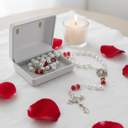 A rosary with white pearl-like beads and red accent beads is displayed beside an open white jewelry box containing a matching rosary. Red rose petals are scattered around on a white cloth, with a lit candle in the background.