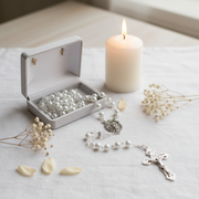 A silver rosary with white pearl-like beads and a silver crucifix is arranged on a white cloth beside an open white jewelry box filled with matching white beads, with a lit white candle and small dried flowers in the background.