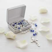 A silver and blue beaded rosary with a Virgin Mary medallion and crucifix, displayed next to an open white jewelry box on a white surface with scattered white rose petals.