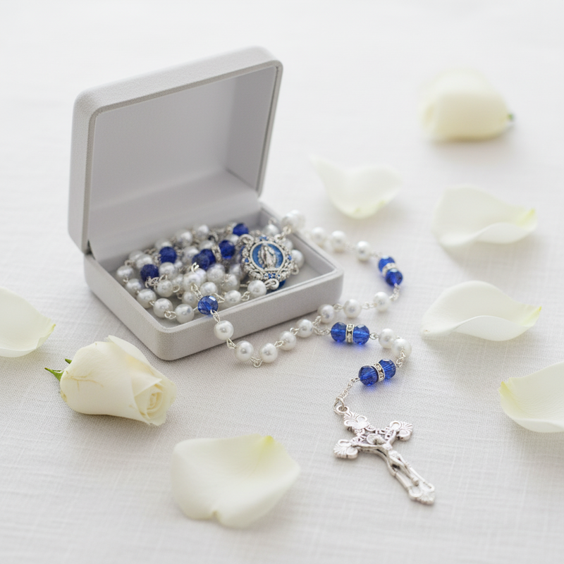 A silver and blue beaded rosary with a Virgin Mary medallion and crucifix, displayed next to an open white jewelry box on a white surface with scattered white rose petals.