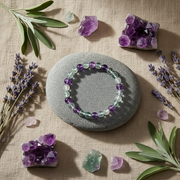 A beaded bracelet made of alternating translucent light green and deep purple beads, displayed on a smooth gray stone. The stone is set against a textured beige linen background and surrounded by decorative natural elements including lavender sprigs, green leaves, and purple crystal clusters.