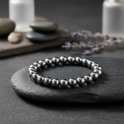 A beaded stretch bracelet made of polished dark metallic beads, displayed on a dark slate stone with a blurred background of candles, smooth stones, and lavender.