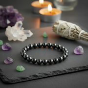 A black beaded bracelet made of polished round beads, displayed on a dark slate tray with purple and green crystals, a white sage bundle, and lit candles in the background.