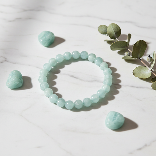 A beaded stretch bracelet made of pale mint-green round stones, arranged on a white marble surface with small mint-green tumbled stones and a sprig of eucalyptus leaves around it.