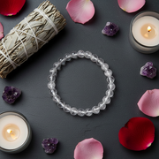 A clear beaded bracelet made of round transparent gemstone beads is arranged in a circle on a dark gray surface. The scene is styled with pink and red flower petals, purple crystal clusters, a bundle of dried grass tied with string, and two lit candles in small glass holders.