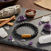 A beaded bracelet made of matte black lava stones with a central yellow-brown carved wooden bead, displayed on a dark slate tray with crystals, a smudge bundle, and a small copper bowl.