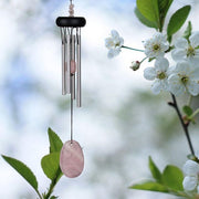 Wind chime with pink stone pendant hanging among white flowers and green leaves.