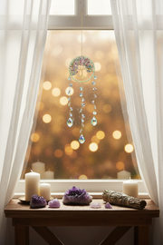 A circular dreamcatcher with hanging crystal beads is suspended in front of a window with white sheer curtains. The window has a warm golden bokeh background, and a wooden table below holds candles, amethyst crystals, and a sage bundle.