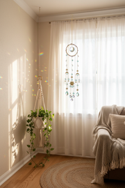 A boho-style dreamcatcher hangs in a sunlit room corner beside sheer white curtains, a hanging plant, and a cozy armchair with a knitted throw blanket.