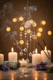 A gold-toned crystal wind chime with pink rose quartz and clear teardrop beads hangs in a mystical setting with burning candles, smudging, and amethyst crystals against a warm bokeh background.
