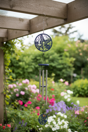 A hanging wind chime with circular blue metal ornaments and silver-toned tubes, suspended under a wooden pergola in a blurred garden setting with flowers and greenery.
