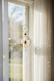 A gold circular window suncatcher with red beads and a clear hanging crystal, suspended in front of a bright window with sunlight and rainbow reflections on the curtains.