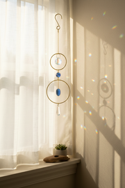 A gold metal suncatcher with blue and clear hanging elements is displayed on a window sill in a sunlit room, casting rainbow light reflections on the wall.