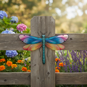 A colorful metal dragonfly garden ornament with vibrant blue, pink, orange, and yellow wings, mounted on a weathered wooden fence in a garden setting.