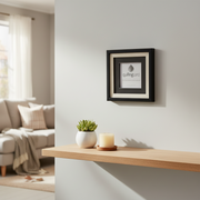 A framed wall art print with a black frame and white mat hangs on a light gray wall above a wooden floating shelf. The print features a gray teardrop logo and the word 'quillingard'.